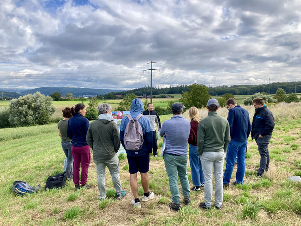 Eine Gruppe Personen steht bei einem Saum auf Ackerland und hört den Ausführungen eines Experten zu